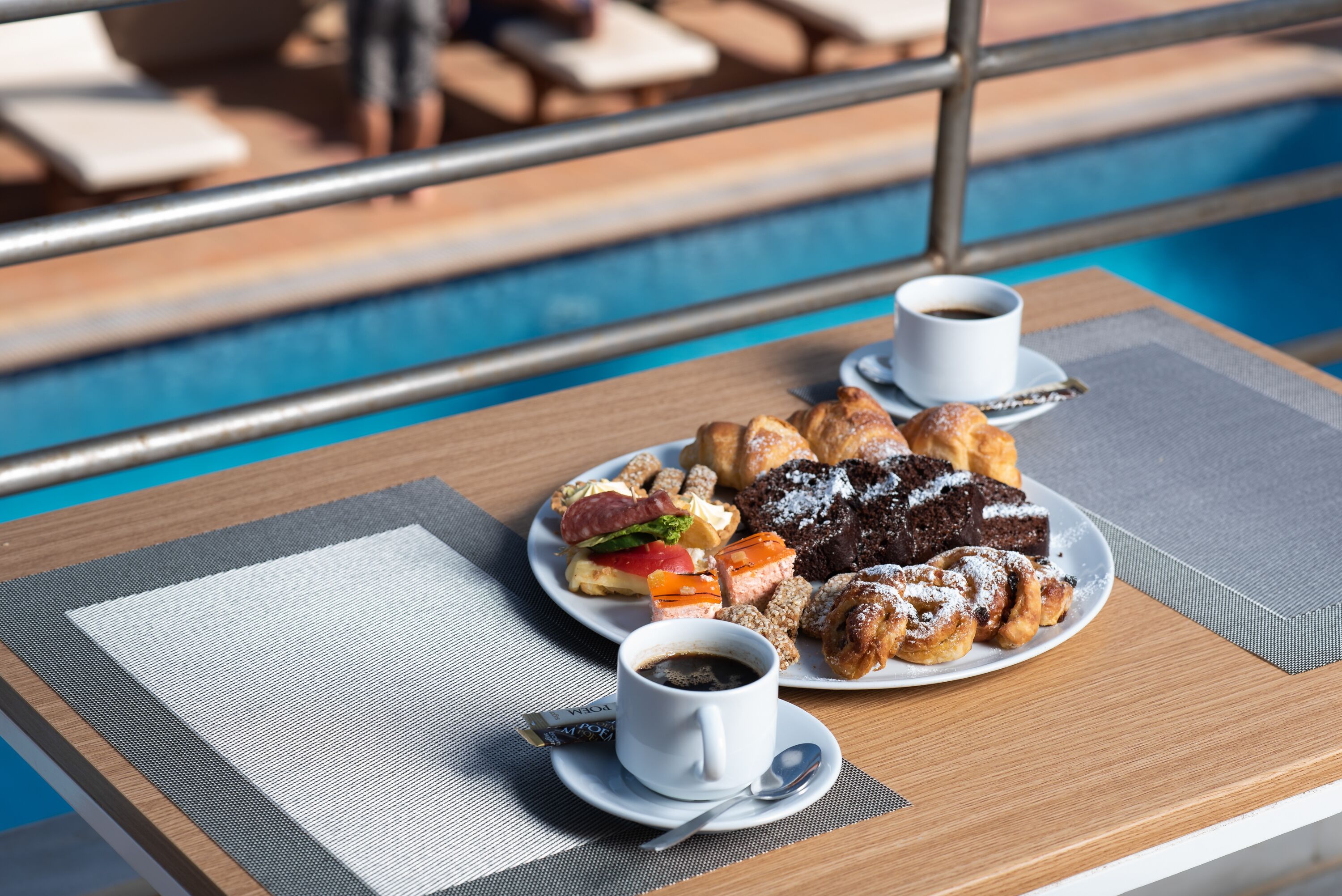 A table with a plate of pastries and two cups of coffee, overlooking a pool area.