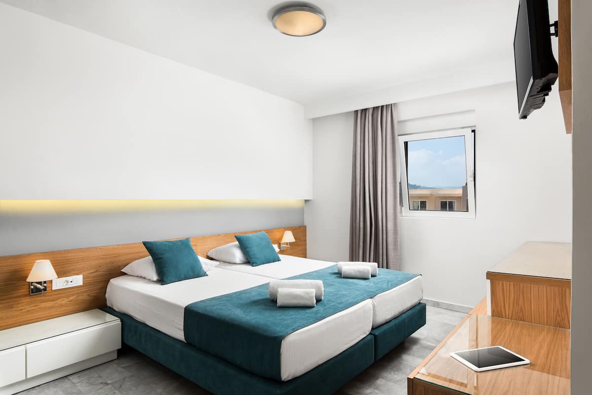 Ett modernt hotellrum med en dubbelsäng, ett nattduksbord med en lampa, en TV och utsikt över stranden genom fönstret.