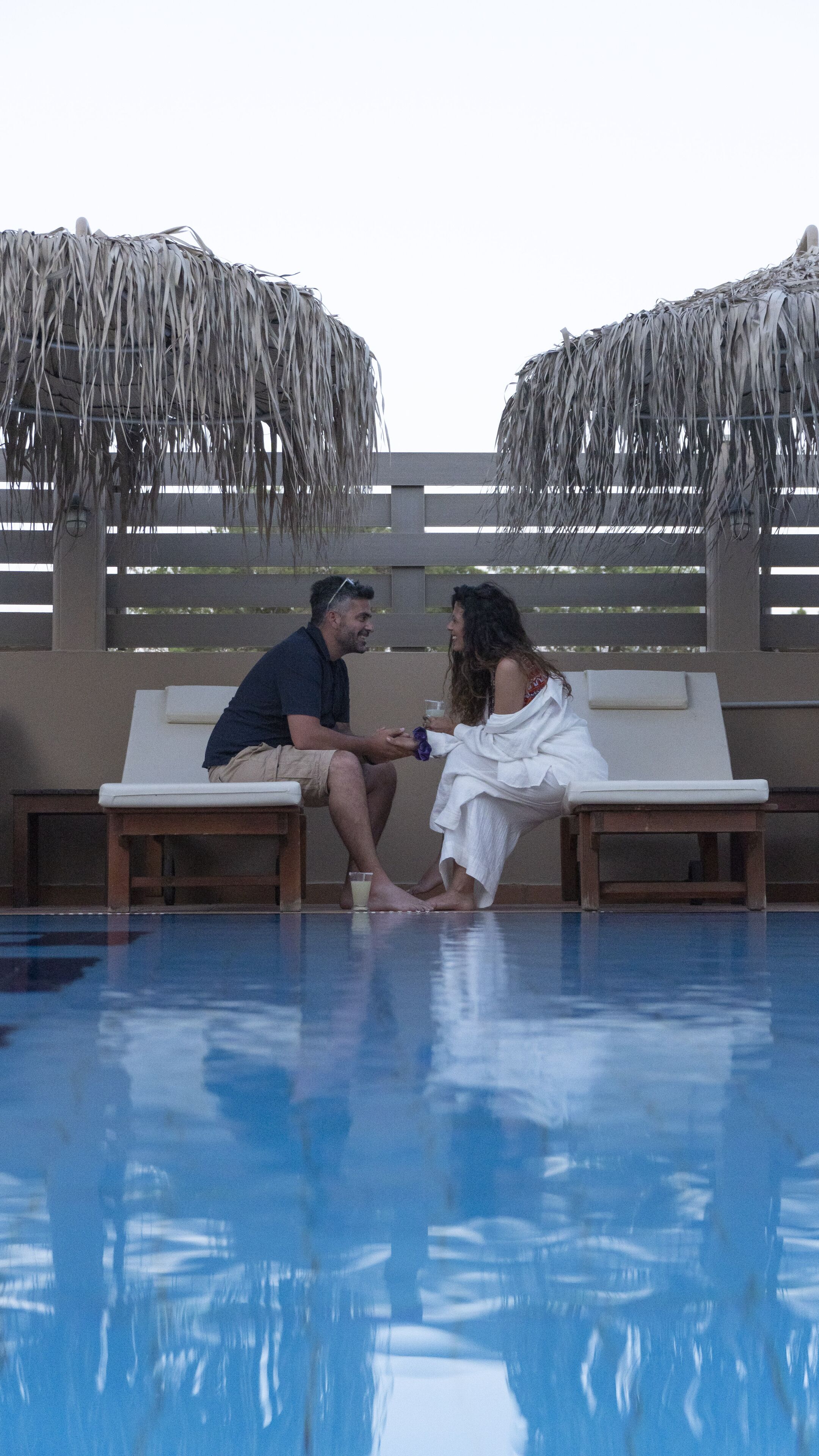 Un couple est assis près d’une chaise longue au bord de la piscine, sous un toit de chaume.