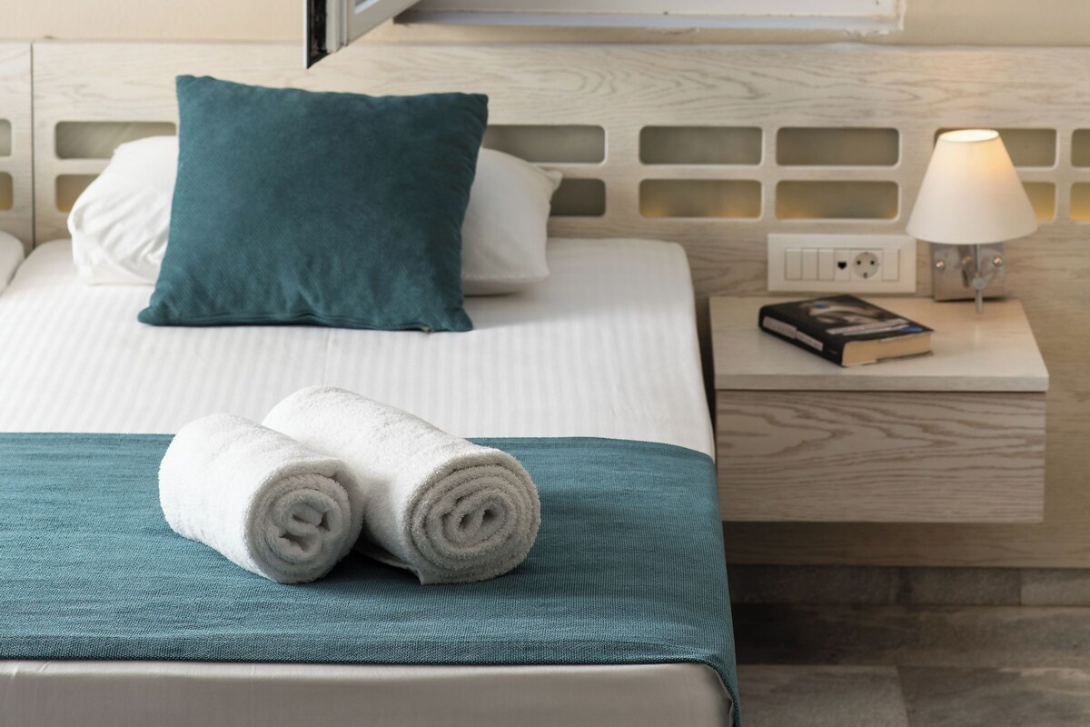 Una habitación de hotel con una cama, un ventanal con vistas a una piscina y al mar, y una mesita de noche con una lámpara y un libro.