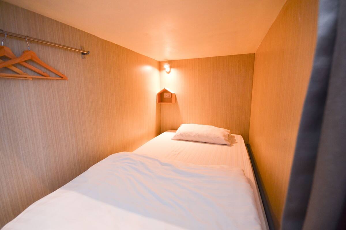 Un petit lit, bien fait, se trouve dans une pièce aux murs en bois, avec une seule lampe suspendue au-dessus.