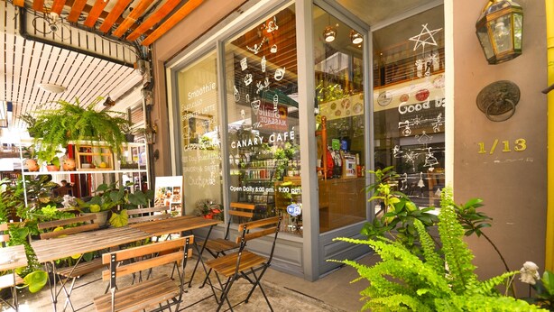 A café with wooden tables and chairs, potted plants, and a glass door with menu items displayed. - Good Day Hostel (Bangkok)
