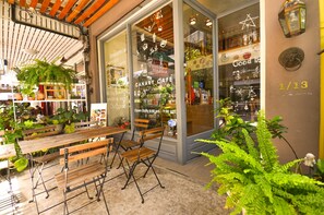 A café with wooden tables and chairs, potted plants, and a glass door with menu items displayed. - Good Day Hostel (Bangkok)