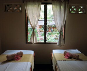 A room with two beds, each with a towel and a hibiscus flower. There are curtains on the window, and a view of greenery outside.