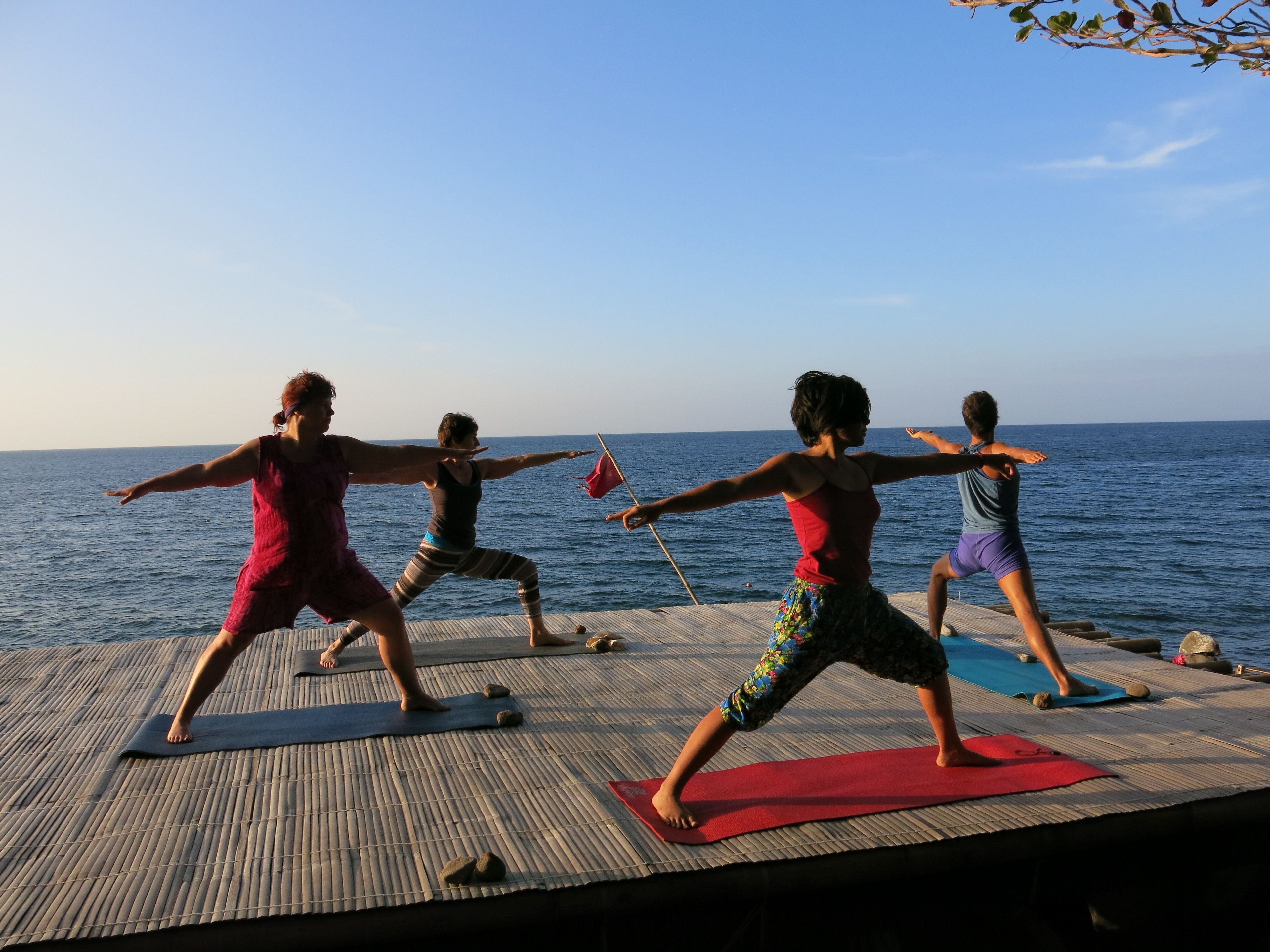 Een groep mensen beoefent yoga op een houten dek, met uitzicht op zee.