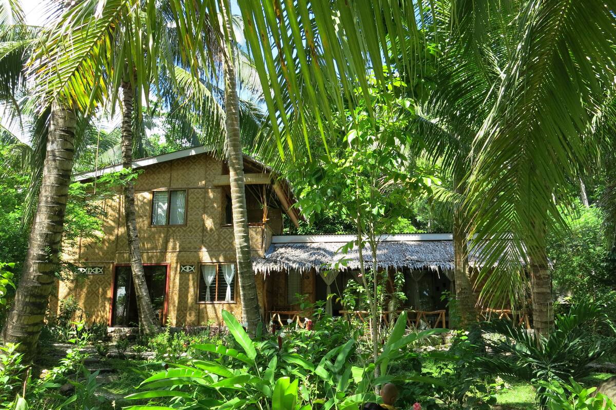 Ein zweistöckiges Haus mit Strohdach, umgeben von üppiger tropischer Vegetation.