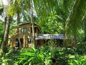 A two-story house with a thatched roof, surrounded by lush tropical vegetation.