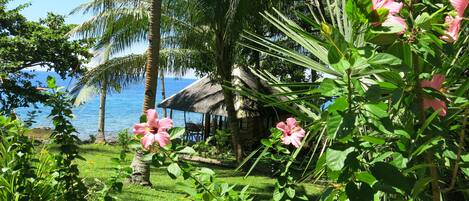 A tropical garden with pink flowers, palm trees, and a thatched-roof structure near a body of water.