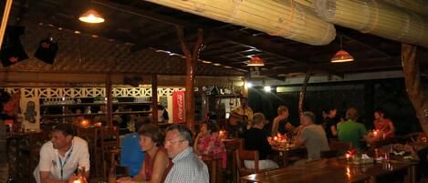 A restaurant with wooden tables and chairs, a bar area with shelves of bottles, and a hanging light fixture.