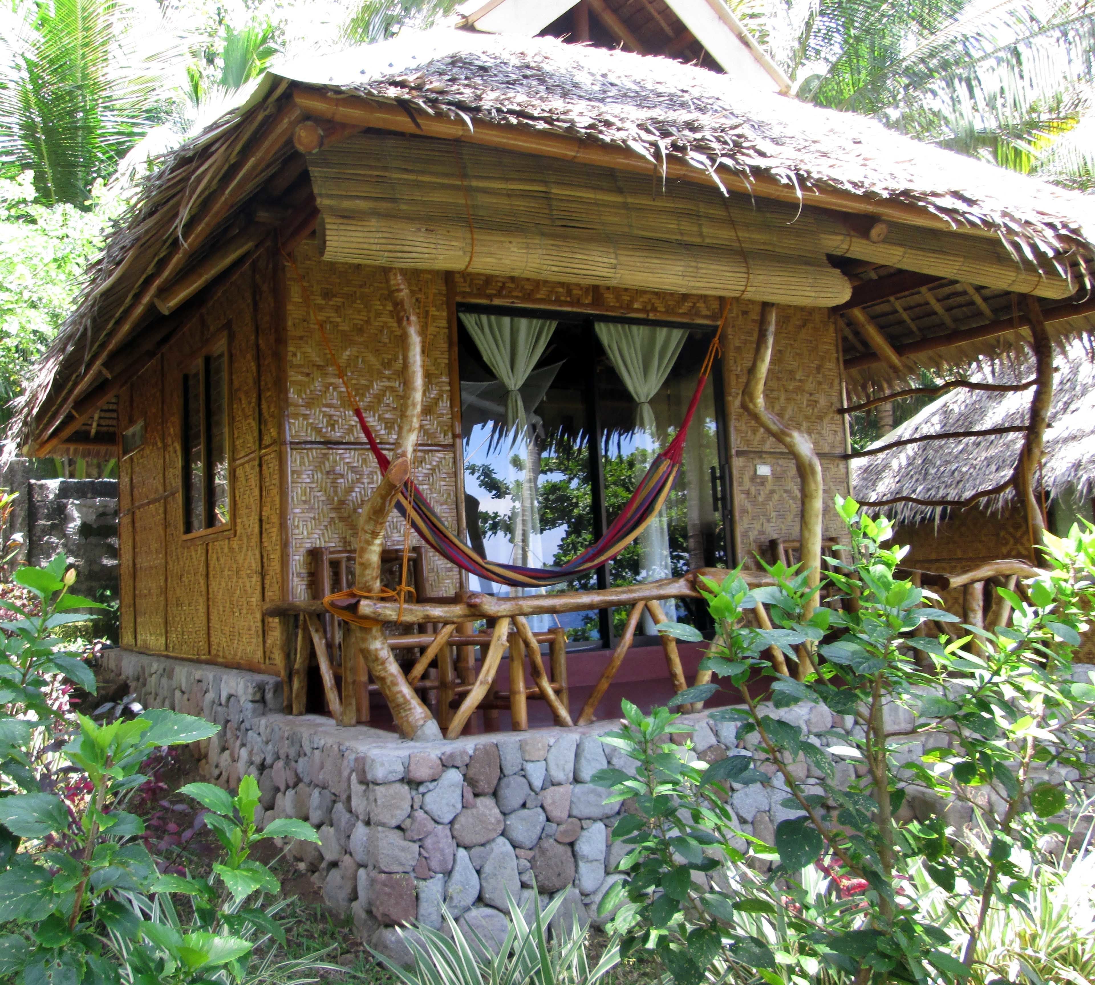 A rustic thatched-roof house with a stone foundation, a hammock, and a window with curtains.
