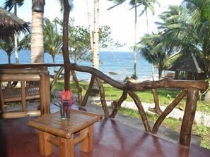 A wooden deck with a table and chairs, overlooking a beach with palm trees and the ocean.