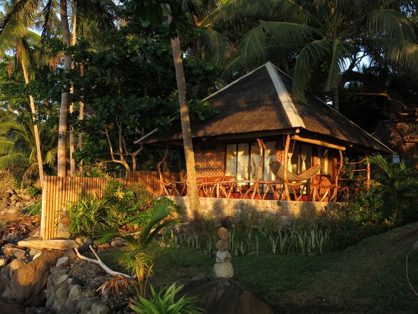 A thatched-roof hut with a wooden deck and a stone wall, surrounded by tropical vegetation and palm trees.