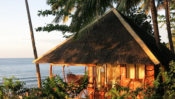 Een rieten bungalow met een houten veranda, omgeven door tropische begroeiing en met uitzicht op de zee.