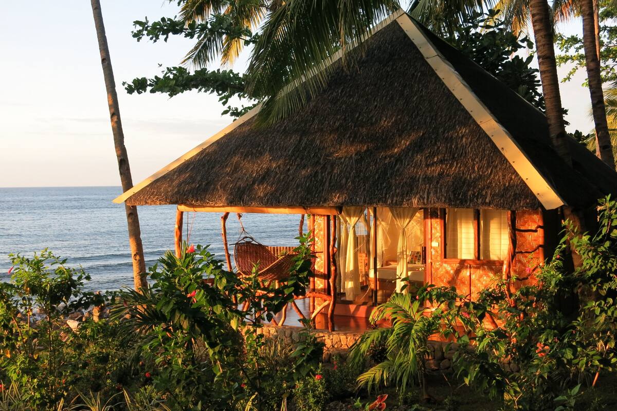 Ein reetgedecktes Bungalow mit einer Holzterrasse, umgeben von tropischer Vegetation mit Blick auf das Meer.