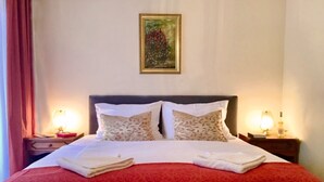 Una cama bien tendida con una colcha roja, dos almohadas y toallas dobladas. Hay dos mesitas de noche con lámparas y un cuadro enmarcado en la pared sobre la cama.