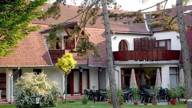 Un edificio de dos plantas con tejado de tejas rojas, un área de estar al aire libre con mesas y sillas, y un pequeño jardín con un árbol y una farola.