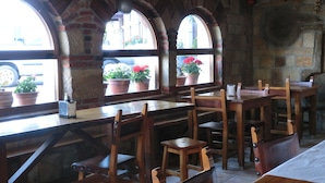 Un interior rústico con mesas y sillas de madera, amplias ventanas de arco con plantas en macetas y una pared de piedra.