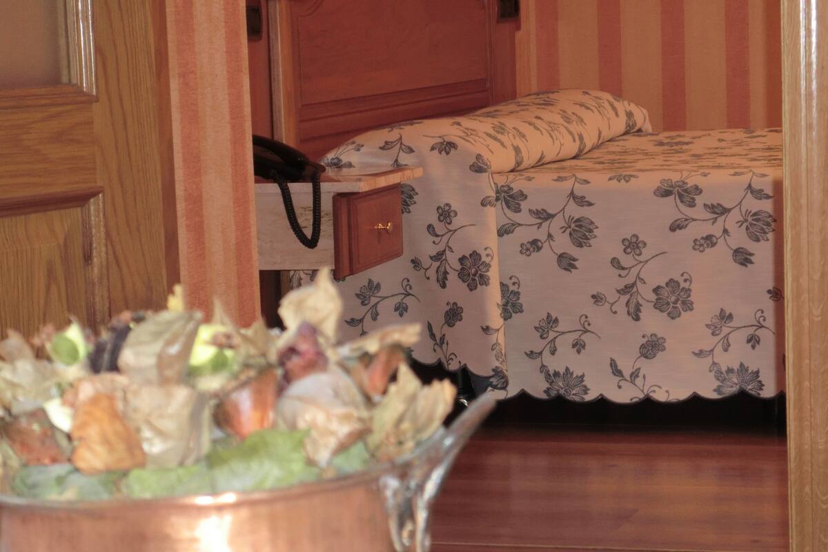 Una habitación con una cama cubierta de ropa de cama estampada con motivos florales, un armario de madera y una olla de cobre llena de verduras.