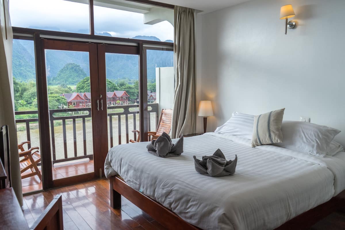 A hotel room with a large bed, a wooden bench, and a view of a mountainous landscape through a glass door.