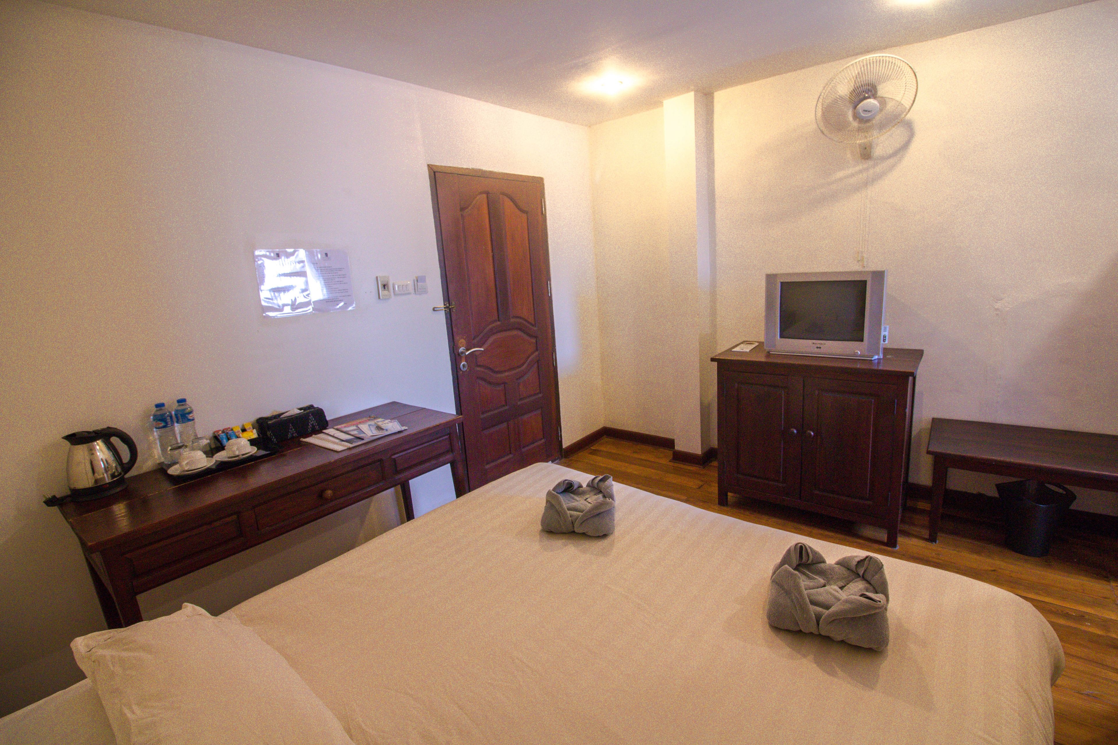 Habitación de hotel con cama, escritorio con tetera, televisor sobre un mueble, armario de madera y ventilador de pared.