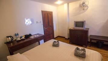 Habitación de hotel con cama, escritorio con tetera, televisor sobre un mueble, armario de madera y ventilador de pared.
