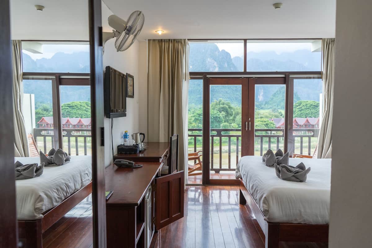 A hotel room with two beds, a desk with a fan, and a view of mountains.