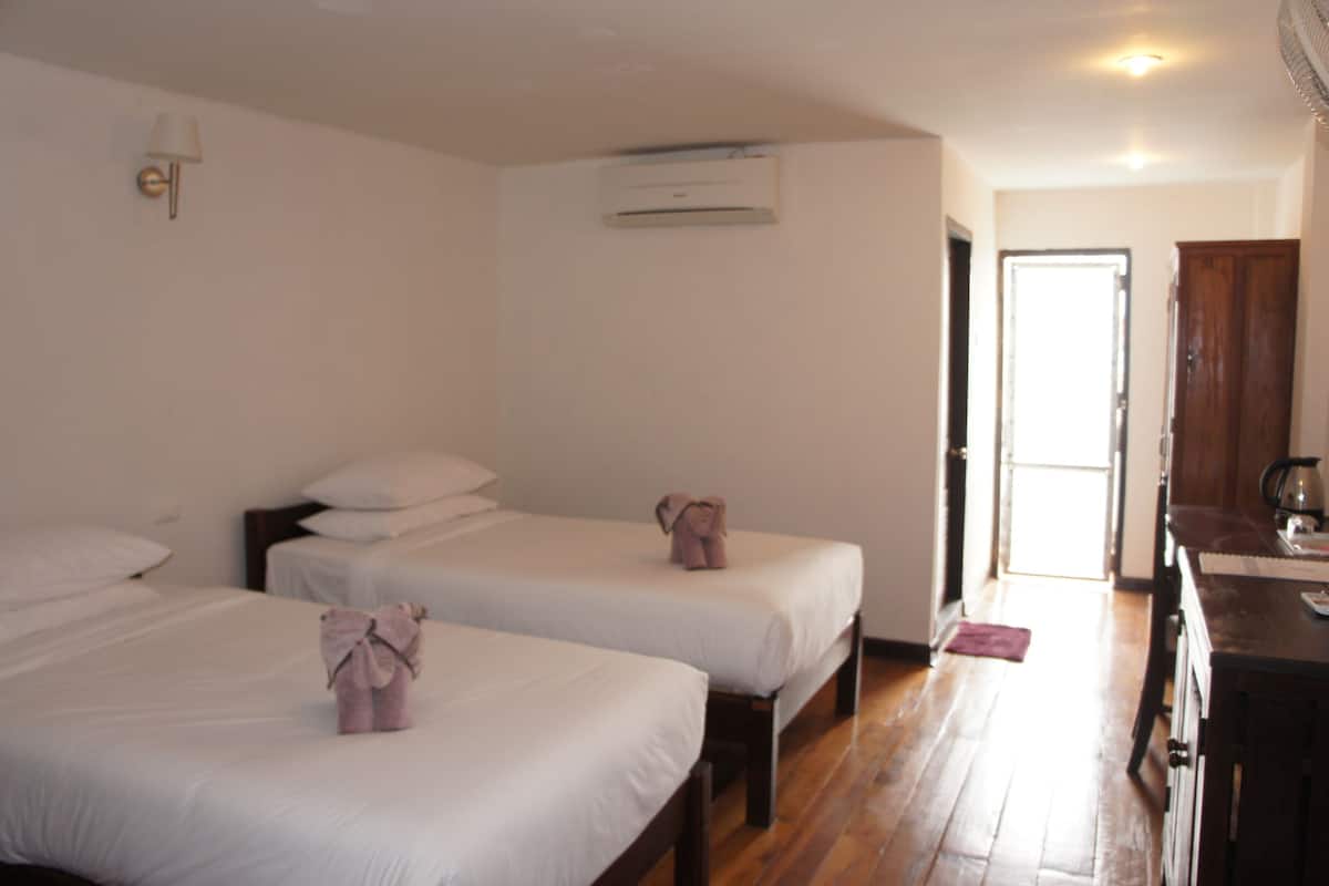 A hotel room with two beds, a wooden floor, an air conditioning unit, a door, and a small table with a kettle.