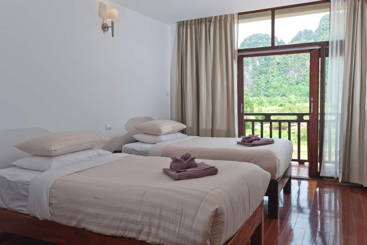 A room with two beds, a wooden floor, a balcony with curtains, and a view of greenery.
