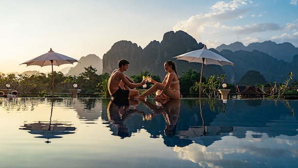 Una pareja sentada junto a una piscina con sombrillas, rodeada de montañas y follaje.