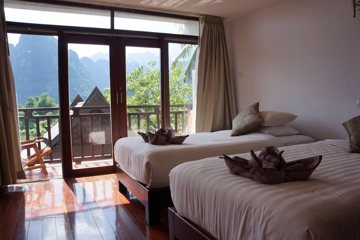 A hotel room with two beds, a balcony with a view of mountains, and a wooden floor.