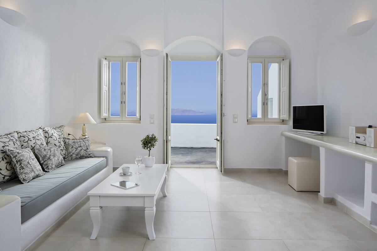 A modern, minimalist room with a white sofa, a coffee table, and a TV on a stand. There are two windows with arched tops, one of which offers a view of the sea.