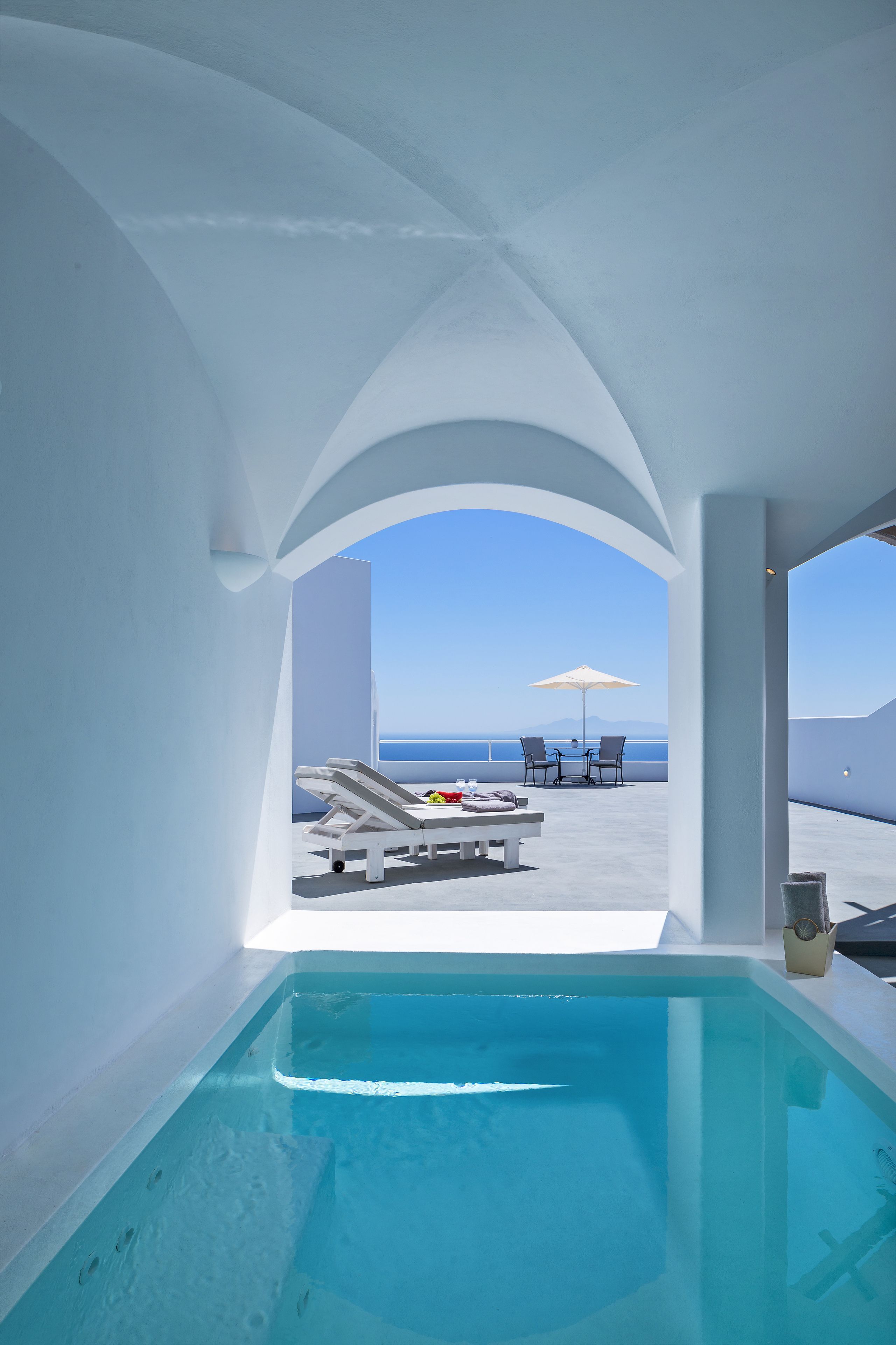 A modern infinity pool with a clear view of the sea, a white interior, and a seating area with umbrellas.