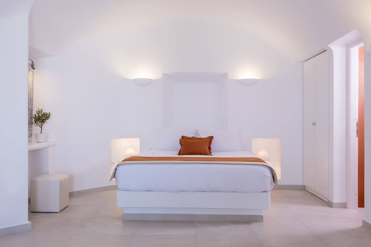 A modern bedroom with a large bed, two bedside lamps, a nightstand, and a potted plant.