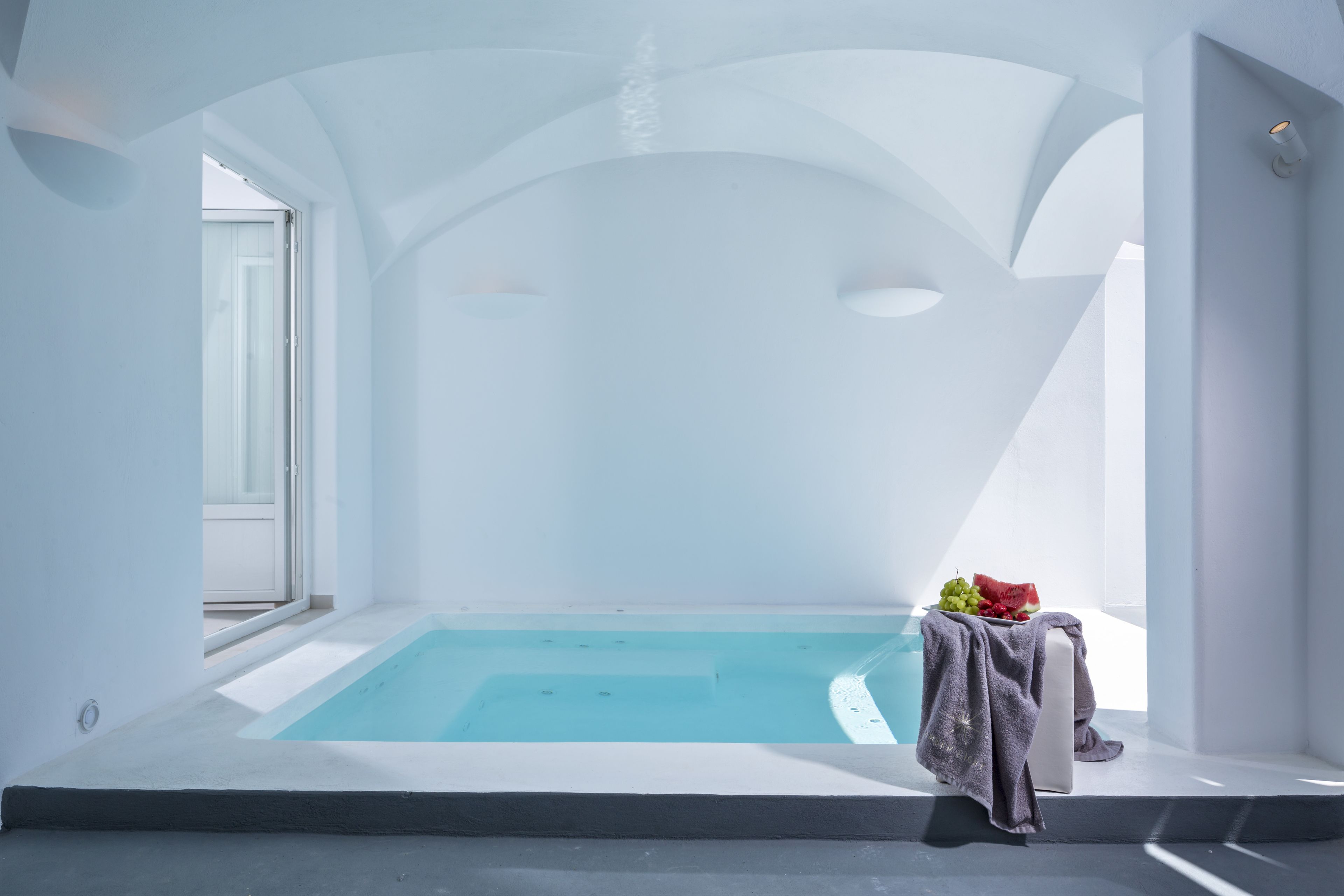 Un área de piscina interior moderna y minimalista con un muro de vidrio transparente, una pequeña mesa con frutas y una toalla sobre un banco.