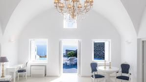 A spacious room with white walls, a high arched ceiling, and a chandelier. There are two windows with views of the sea, a white table with chairs, and a sofa.
