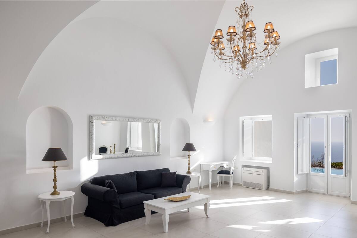 A spacious living room with a dark sofa, a white coffee table, and a chandelier. There is a white desk with a chair and a large mirror on the wall.