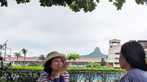 Zwei Personen sitzen an einem Tisch im Freien mit Blick auf ein Gebäude und einen Berg im Hintergrund.