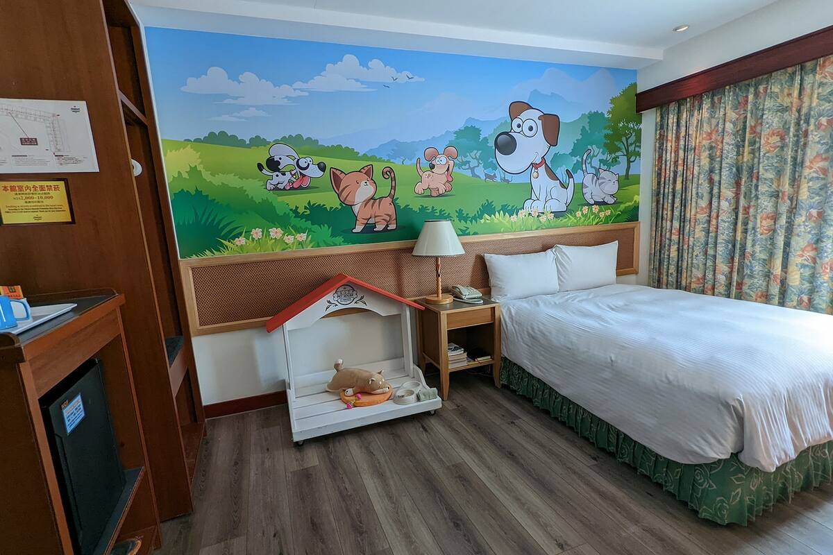 Ein Hotelzimmer mit einem Bett, einem Nachttisch, einer Lampe, einem kleinen Tisch mit einer Katzenfigur und einem Wandgemälde mit Zeichentieren.