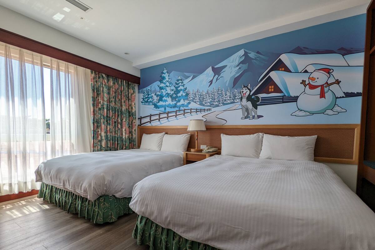 Ein Hotelzimmer mit zwei Betten, einem hölzernen Kopfteil, einem Nachttisch mit Lampe und einem Wandbild mit einer verschneiten Berglandschaft.