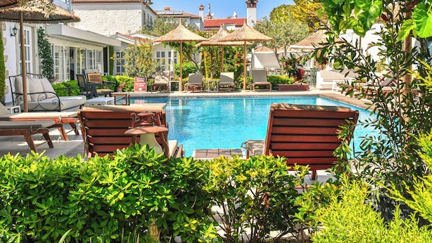 A pool area with lounge chairs and umbrellas, surrounded by greenery and buildings with red-tiled roofs. - Bay C Hotel (Cesme)