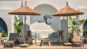A poolside area with lounge chairs, two thatched umbrellas, and a wall with the logo "BAY C". - Bay C Hotel (Cesme)