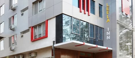 Un hotel moderno con una fachada destacada en rojo y gris, varios balcones y una entrada bien cuidada con plantas en macetas.