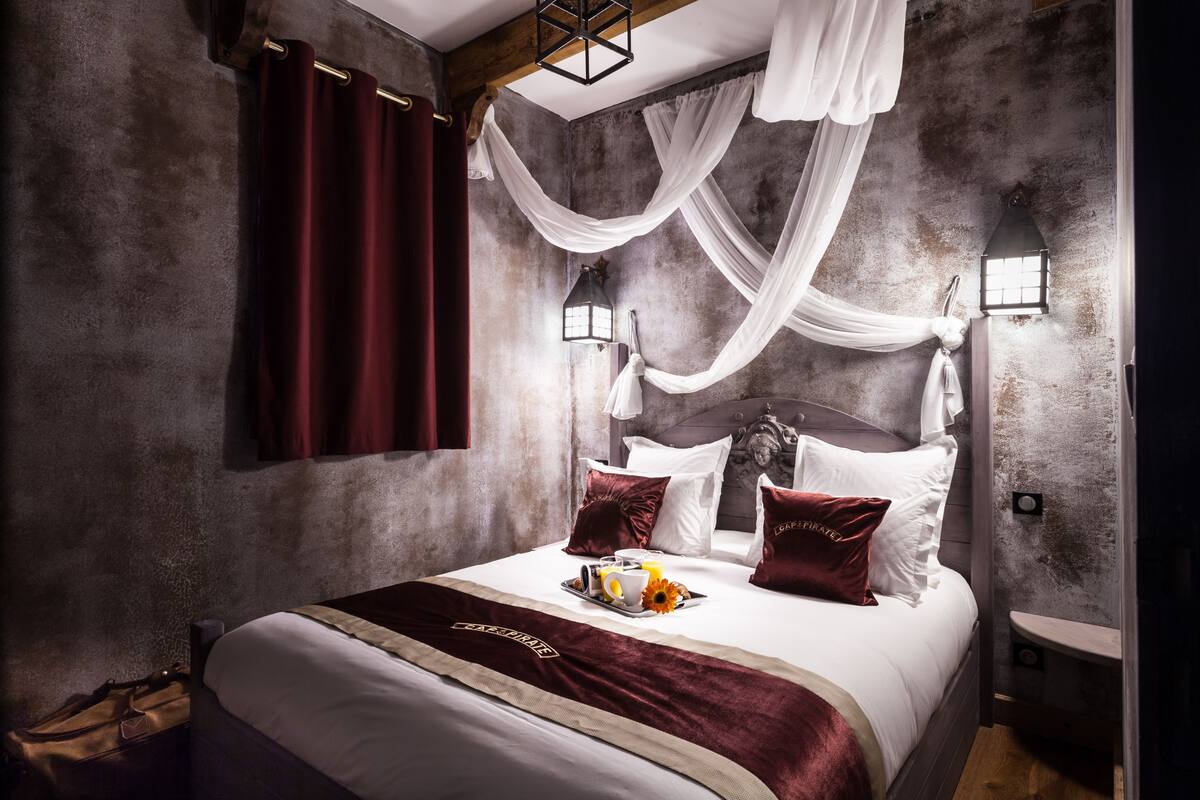 A bedroom with a large bed, draped white curtains, a headboard with a decorative headpiece, and a tray of breakfast items on the bed.