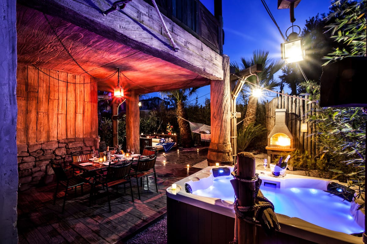 An outdoor patio with a hot tub, wooden tables, and chairs, surrounded by stone walls and plants.