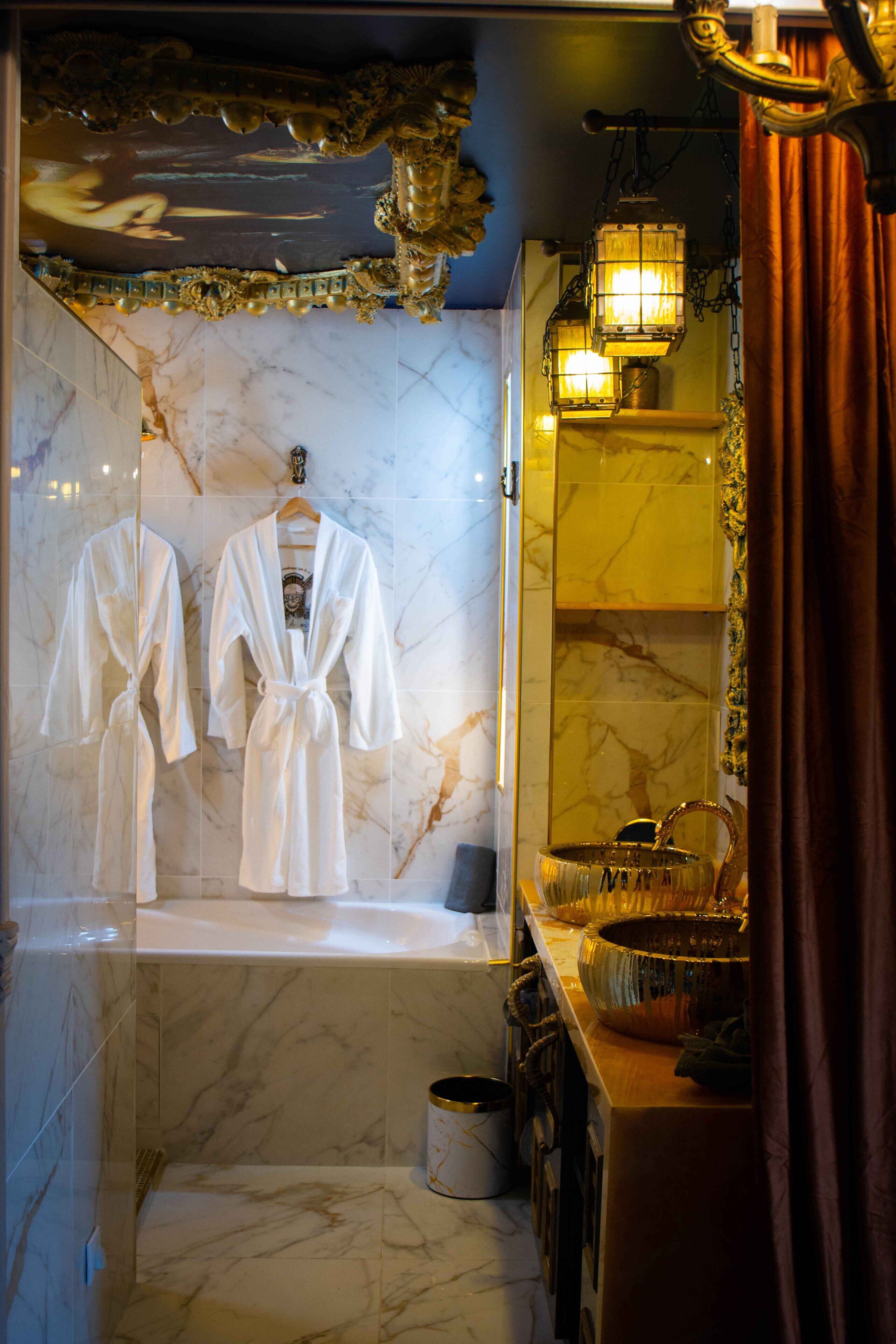 A bathroom with marble walls, a bathtub, a sink with a mirror, and two white robes hanging on a rack.