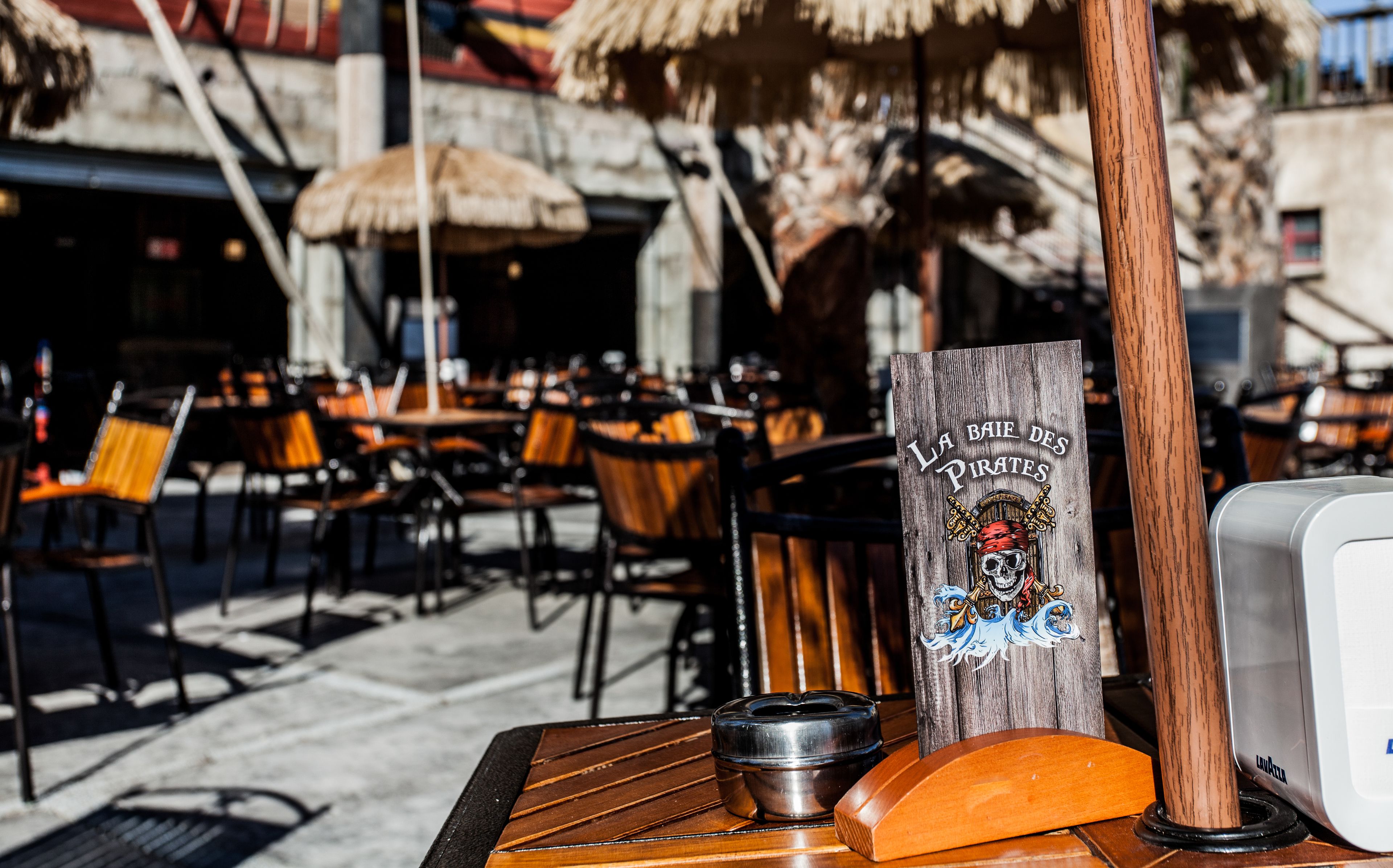 An outdoor seating area with wooden tables and chairs, a wooden sign with a pirate theme, and a thatched umbrella.