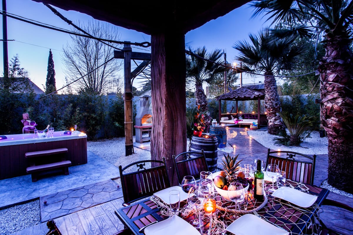 An outdoor patio with a dining table set for two, surrounded by chairs, a hot tub, and a fire pit.