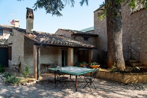 Un patio rústico al aire libre con una mesa y sillas, rodeado de edificios de piedra y un árbol grande.