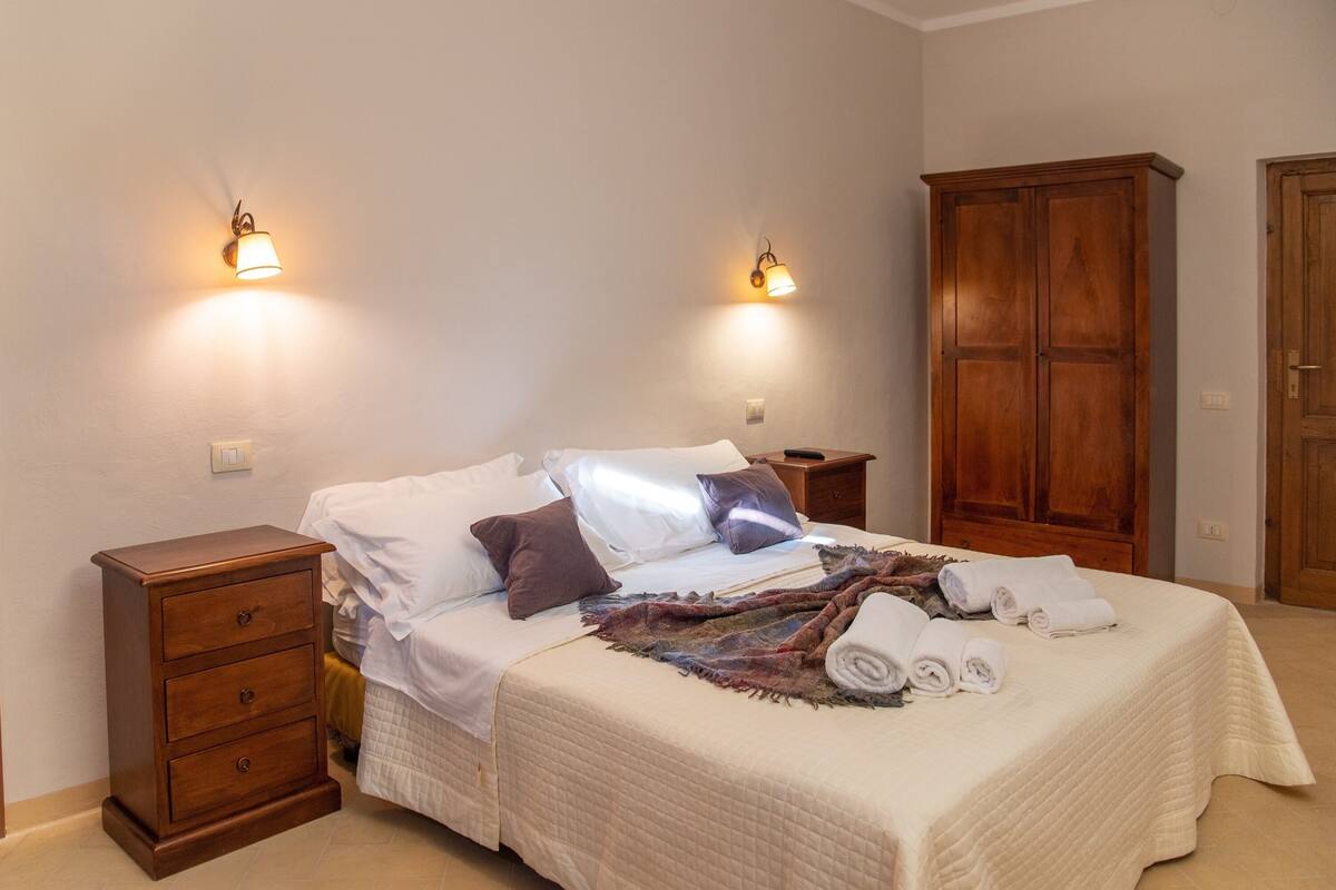 Een opgemaakt bed met kussens en dekbed, een nachtkastje met lades, en een kledingkast.
