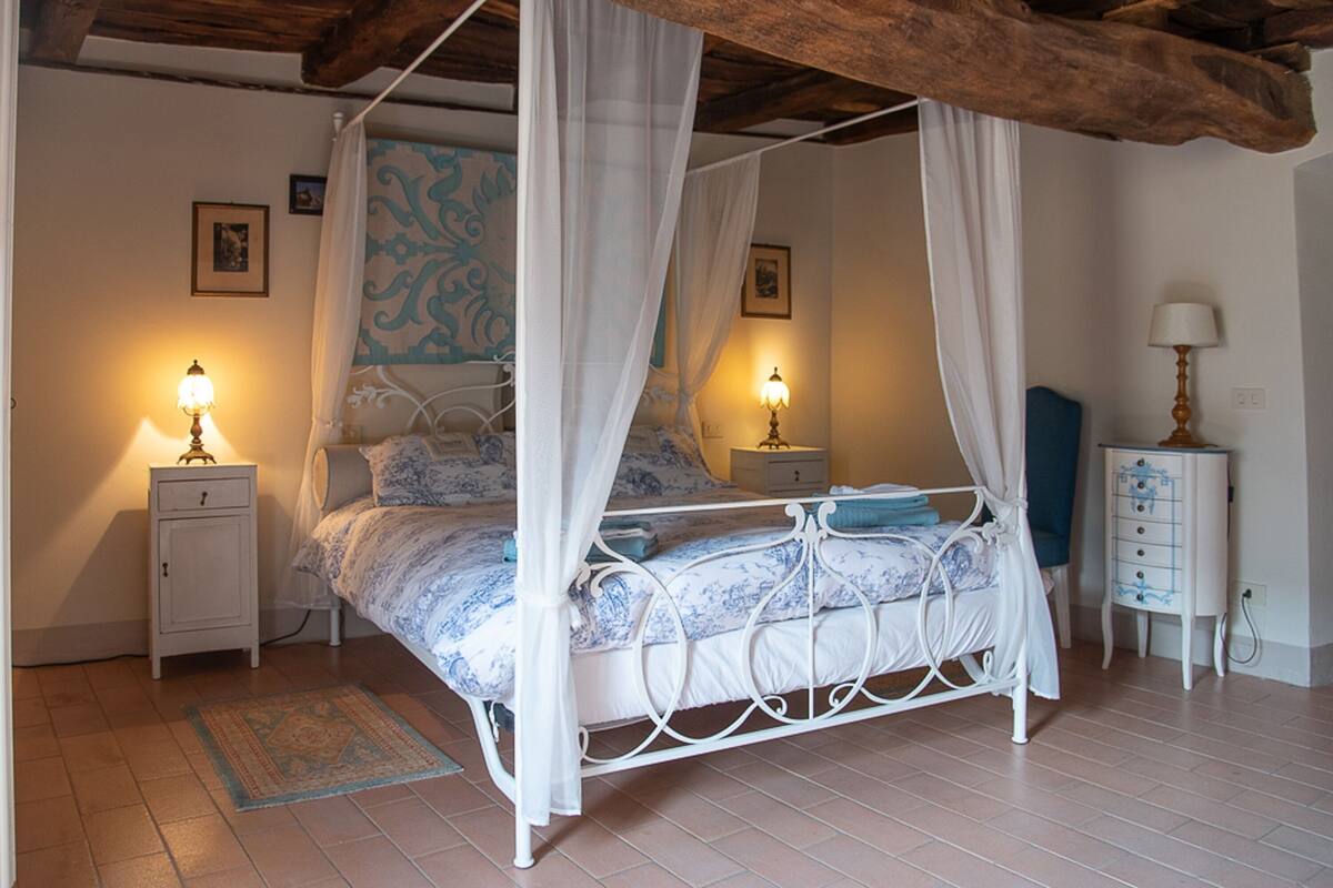 Een hemelbed met hemeltje, twee nachtkastjes met lampen, een klein dressoir en een kamerbreed tapijt met dessin.